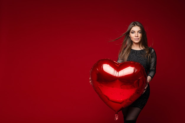 Free photo portrait of stunning brunette girl in dark cocktail dress with red heart balloon on red background. saint valentines day concept.