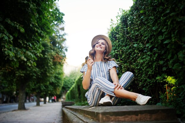 Portrait of a stunning young woman in striped overall sitting in the park and listening to the music with her earphones on