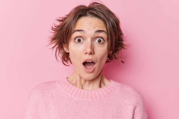 Portrait of stupefied emotional young woman stares at camera keeps mouth widely opened screams in panic looks with terror has trendy hairstyle wears casual jumper isolated over pink background