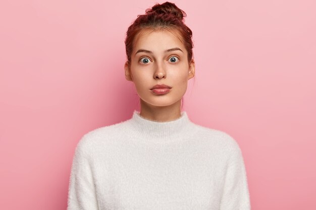 Portrait of stupefied young dark haired woman with hair, dressed in casual white sweater, poses against pink background.