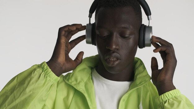 Portrait of stylish African American guy wearing bright jacket listening music in headphones isolated on white background. Cool boy