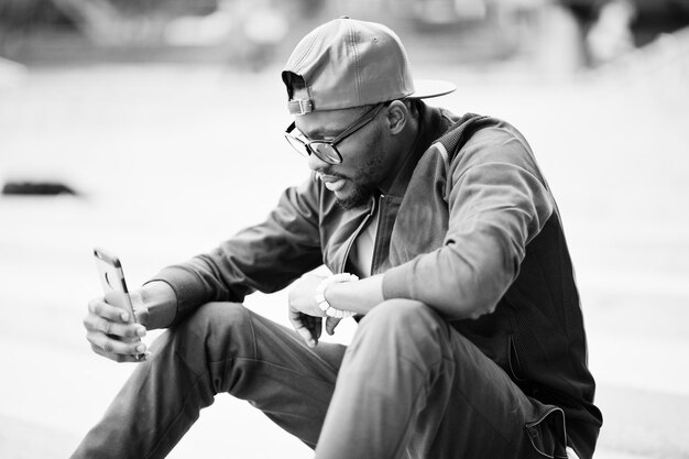 Portrait of stylish african american man on sportswear cap and glasses sitting on stairs with phone at hand Black men model street fashion