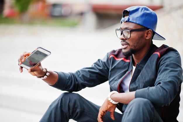 Portrait of stylish african american man on sportswear cap and glasses sitting on stairs with phone at hand Black men model street fashion