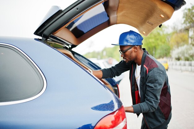 Portrait of stylish african american man on sportswear cap and glasses walking with handbag and open car trunk Black men model street fashion