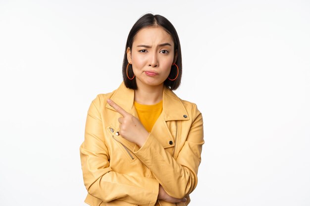 흰색 배경에 서 있는 로고나 광고에 손가락을 왼쪽으로 가리키는 의심스러운 얼굴로 바라보는 세련된 아시아 소녀의 초상화