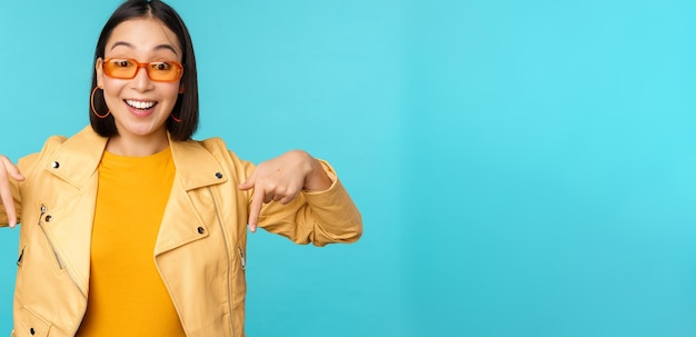Портрет стильной азиатской девушки в солнцезащитных очках улыбается и указывает пальцем вниз, показывая рекламные стенды на синем фоне