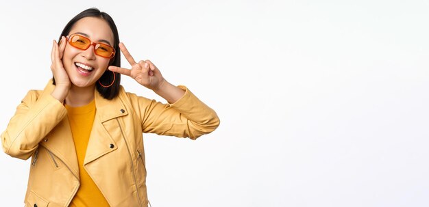 Ritratto di elegante ragazza asiatica moderna che indossa occhiali da sole e giacca gialla che mostra il gesto di pace vsign in piedi su sfondo bianco faccia sorridente felice