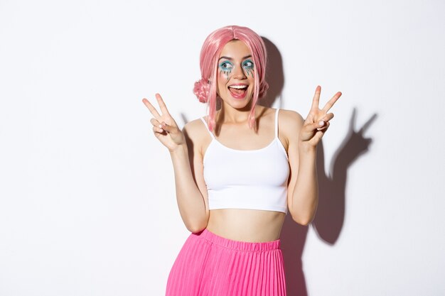 Portrait of stylish attractive girl in pink wig and halloween costume, showing peace signs and smiling happy, standing.