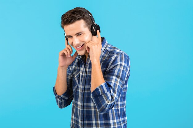 Portrait of stylish attractive handsome young man listening to music on wireless headphones having fun modern style happy emotional mood isolated on blue background wearing checkered shirt