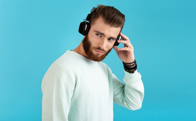 Portrait of stylish attractive young bearded man listening to music on wireless headphones modern style confident mood isolated on blue background