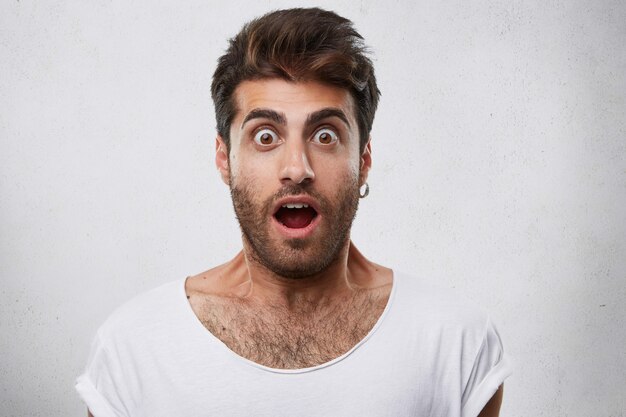 Portrait of stylish bearded guy having trendy hairstyle wearing earring and white T-shirt looking with his eyes popped out and opened mouth having shock and scared look.