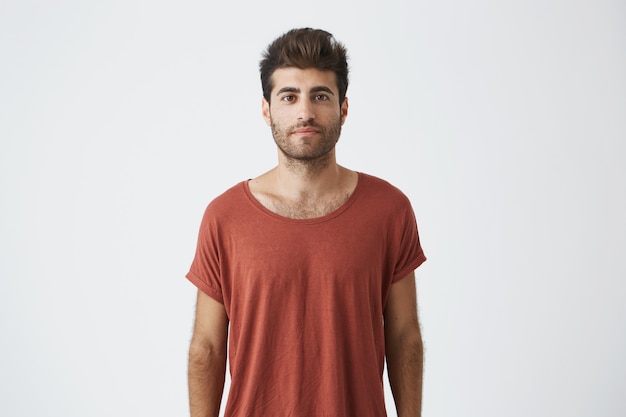 Portrait of stylish bearded guy with trendy haircut wearing casual red T-shirt looking with his brown eyes . Young handsome man having pleased look. People and emotions concept