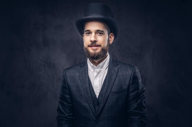 Portrait of a stylish bearded male in an elegant suit and cylinder hat, looking at a camera over dark background.