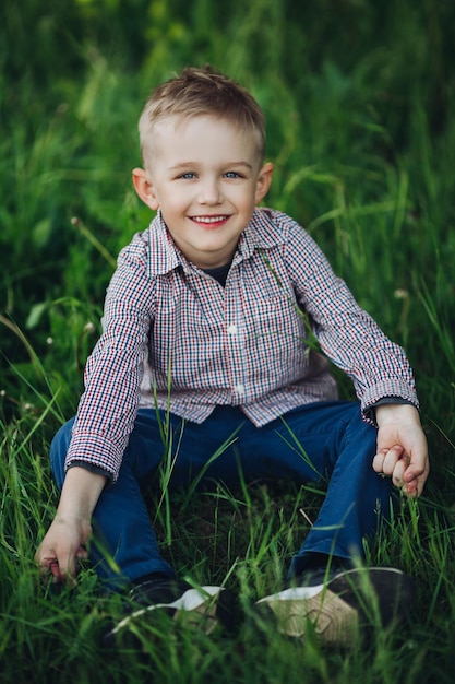 Portrait of stylish blonde little boy wearing in checkered shirt and jeans sitting in park at summertime among grass Handsome boy playing and smiling at camera Kids look and fashion