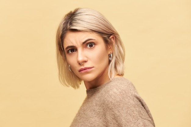 Portrait of stylish blonde young woman wearing cozy sweater staring in indignation, frowning eyebrows, being disgusted with bad smell, stink. Indignant girl expressing displeasure