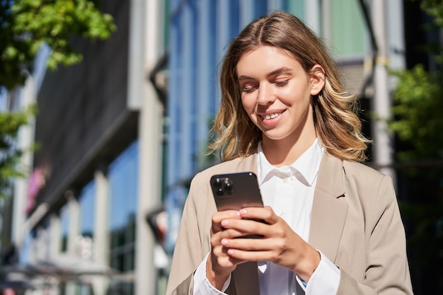 Portrait of stylish businesswoman in corporate outfit holding smartphone standing on street and usin