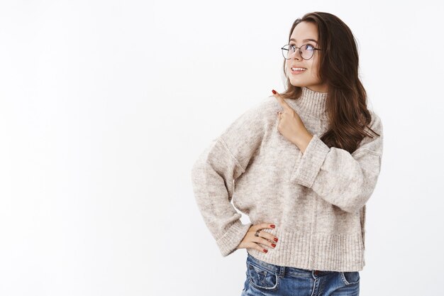 Portrait of stylish and confident good-looking female freelancer in glasses and sweater looking and pointing at upper left corner with pleased intrigued smile observing interesting copy space.