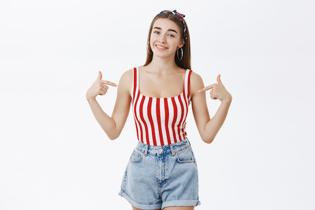 Portrait of stylish confident and proud european woman in trendy striped top and shorts pointing at herself with satisfied happy smile being assertive and self-assured posing against white wall