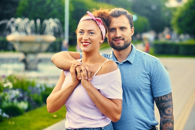 Free photo portrait of stylish couple over city fountain. a man with tattoos on his arms is hanging a redheaded woman.