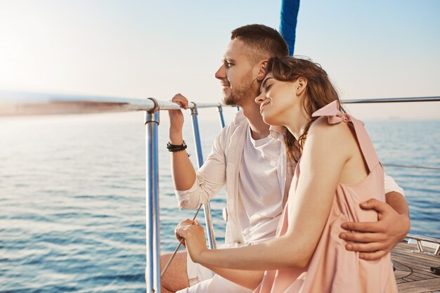 Portrait of stylish couple in love, hugging while sitting on bow of private yacht and enjoying view of sea. Husband took his wife to beautiful warm country, celebrating honeymoon