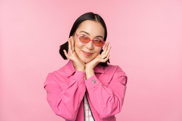 Portrait of stylish cute asian girl smiling and touching face looking up dreamy thoughtful look standing over pink background