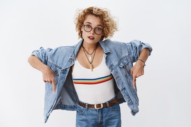 Portrait of stylish european female in glasses and denim looking confused and questioned at front pointing down with index fingers as asking question about unfamiliar product over white wall