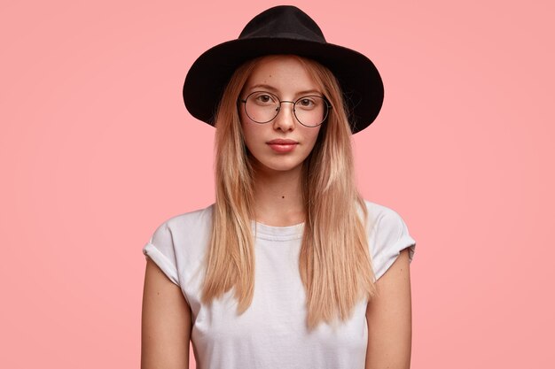 Portrait of stylish European woman wears spectacles and fashionable black hat, has serious expression