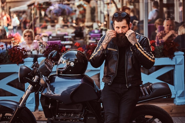 Портрет стильного модного байкера, одетого в черную кожаную куртку, поправляющего усы, сидящего на своем сделанном на заказ ретро-мотоцикле возле террасы кафе.