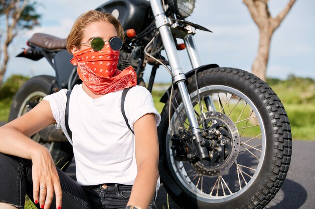 Portrait of stylish female biker wears bandana and sunglasses, sits near fast motorbike, looks pensively away, has rest in open air after long ride, enjoys freedom and high speed. Hobby concept