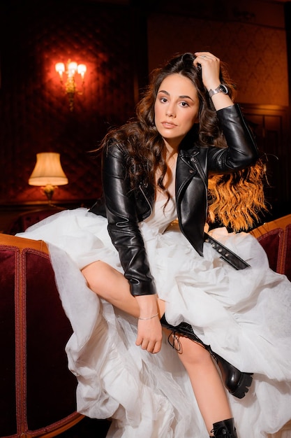 Portrait of stylish female model with curly long hair wearing in white wedding dress black leather jacket and boots sitting on couch in dark room seriously posing and looking at camera