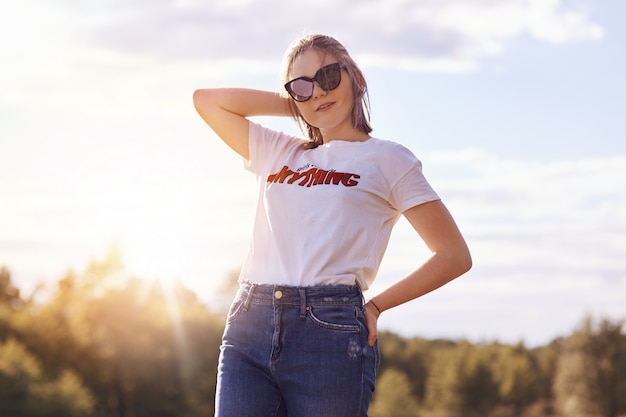 Portrait of stylish female youngster in casual clothes and shades, enjoys summer time, poses on clear sky, has trendy hairstyle. Teenage girl admires beautiful nature. People, style, friendship