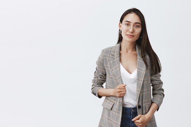 Portrait of stylish feminine woman in trendy jacket and glasses, smiling politely and standing over gray wall