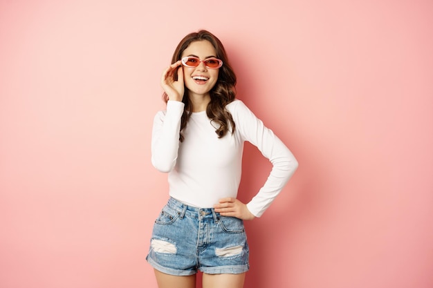 Portrait of stylish glam girl in sunglasses laughing and smiling standing over holiday pink background