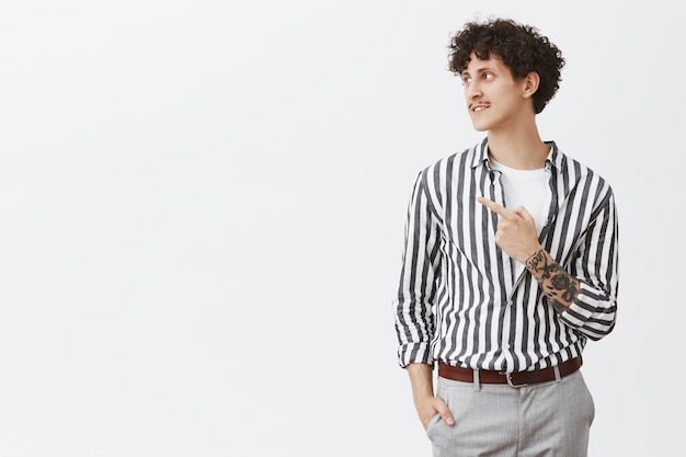 Portrait of stylish good-looking and confident modern hipster male model with moustache curly dark hair and tattoos pointing and gazing left with pleased charming smile over gray wall