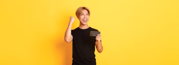 Портрет стильного красивого азиатского парня, играющего в игру на смартфоне и радующегося победе в стоячем желтом