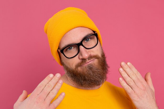Портрет стильного красивого европейского бородатого мужчины в повседневной желтой рубашке, шляпе и очках на розовом фоне