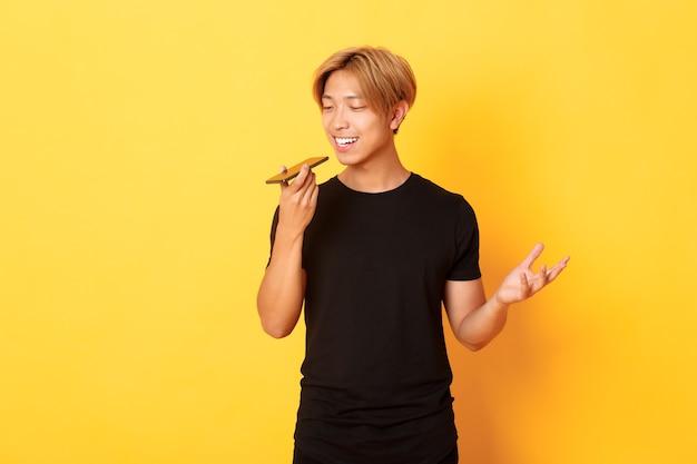 Portrait of stylish handsome korean guy with blond hair record voice message on mobile phone, holding smartphone near mouth and speaking, standing yellow wall