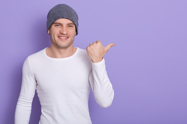 Portrait of stylish handsome young man osing isolated on lilac. Male smiling and points aside, wears white casual long sleeve shirt and gray cap. Copy space for advertisment or promotion.