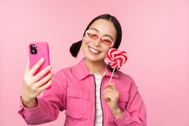 キャンディーロリポップのお菓子で自分撮りを撮って、ピンクの背景の上に立っているモバイルアプリで写真を撮って笑っているスタイリッシュな幸せなアジアの女の子の肖像画