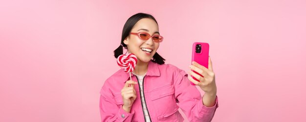 Portrait of stylish happy asian girl taking selfie with candy lolipop sweets and smiling taking photo with mobile app standing over pink background