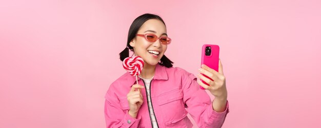 キャンディーロリポップのお菓子で自分撮りを撮って、ピンクの背景の上に立っているモバイルアプリで写真を撮って笑っているスタイリッシュな幸せなアジアの女の子の肖像画