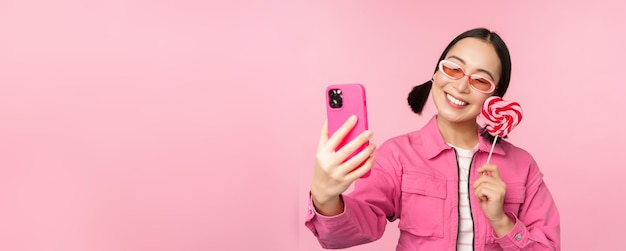 キャンディーロリポップのお菓子で自分撮りを撮って、ピンクの背景の上に立っているモバイルアプリで写真を撮って笑っているスタイリッシュな幸せなアジアの女の子の肖像画