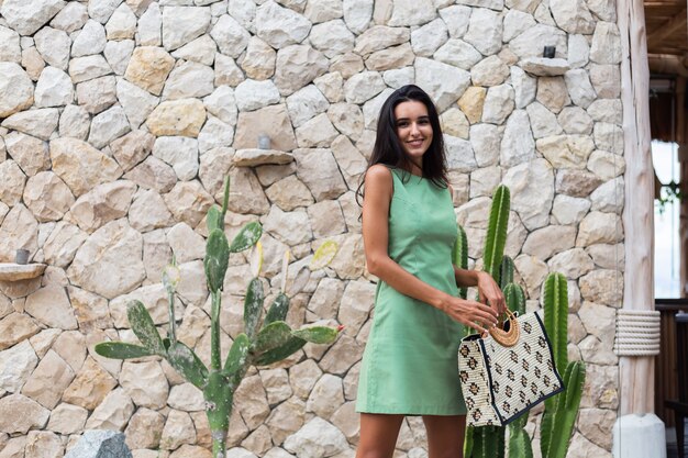 Portrait of stylish happy cute smiling woman in elegant summer green dress holding bag wearing straw hat on background of white stone wall and cactus