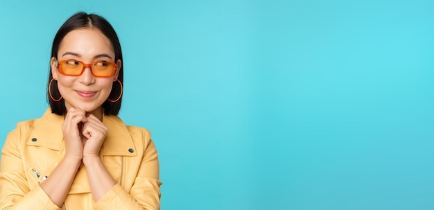 Portrait of stylish korean woman in sunglasses smiling and looking aside at logo coquettish standing over blue background