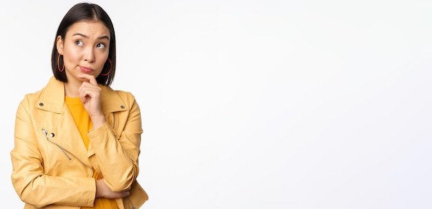 Portrait of stylish korean woman in yellow jacket smiling thoughtful thinking and looking up at logo or advertisement standing over white background