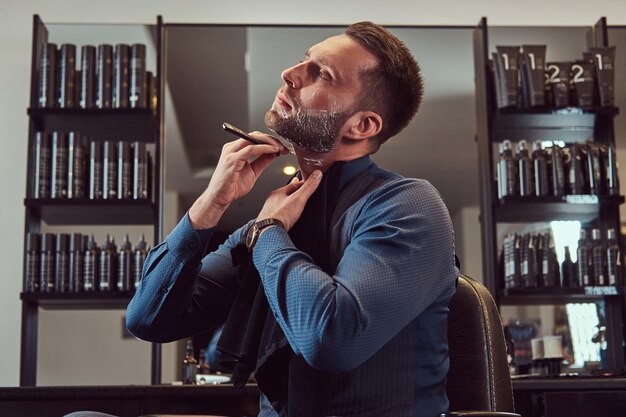 Portrait of a stylish male who himself shaving while sitting on a barber chair in a hairdressing salon.