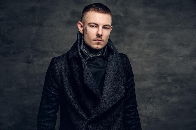 Portrait of a stylish male with tattoo on his face, dressed in a jacket over grey background.