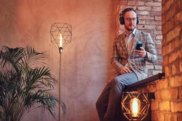 ロフトのインテリアの部屋の窓枠に座って電話とヘッドフォンで音楽を聴いているフランネルのスーツとメガネのスタイリッシュな男の肖像画。