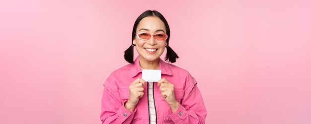 Portrait of stylish modern asian girl shows discount credit card and look pleased paying contactless concept of shopping standing over pink background copy space