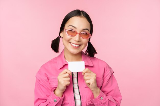 Free photo portrait of stylish modern asian girl shows discount credit card and look pleased paying contactless concept of shopping standing over pink background